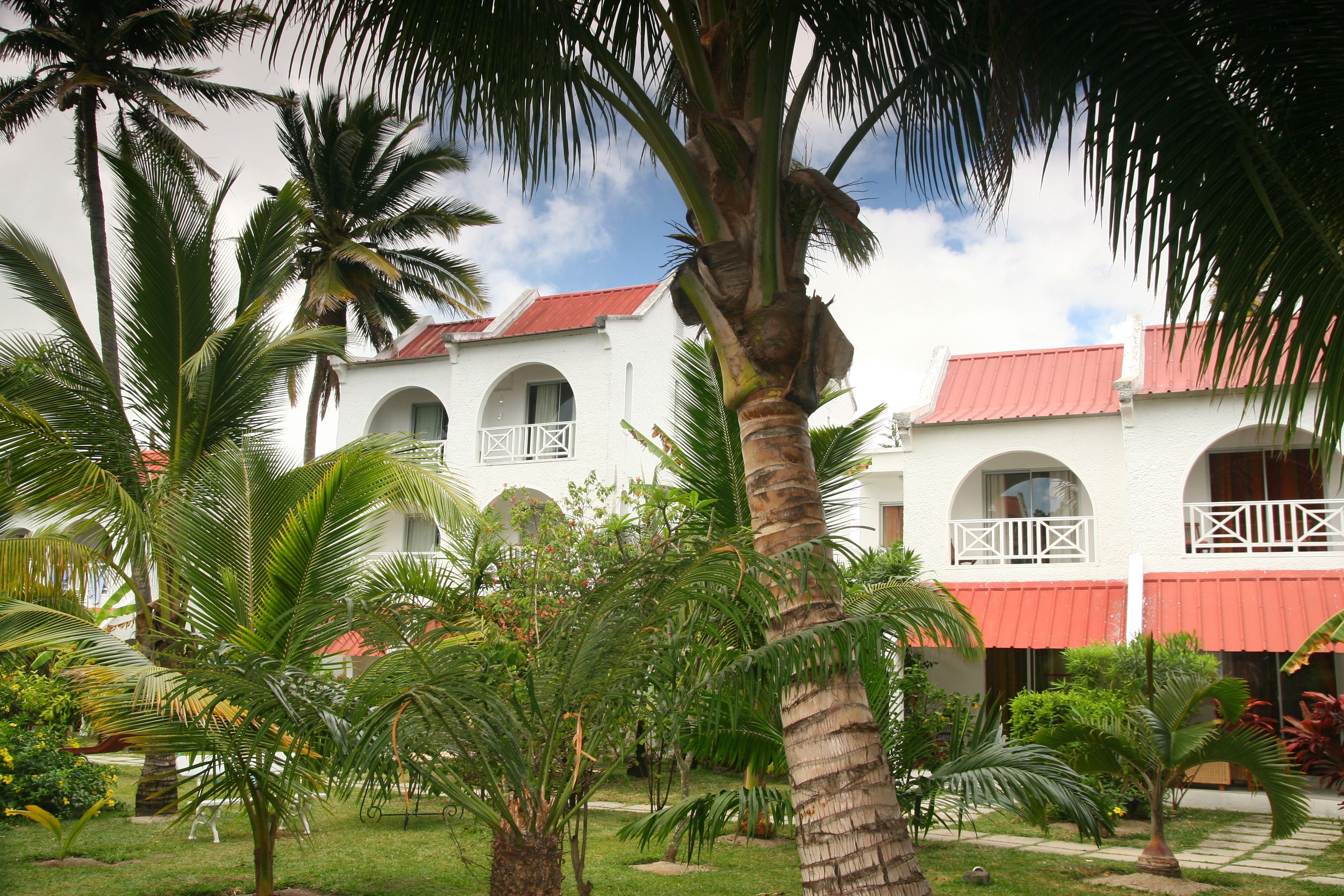 A white building with red roofs and arched windows, surrounded by palm trees and greenery.