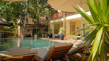 Un espace aménagé au bord de la piscine, avec des chaises longues et des parasols, entouré de plantes en pot et bordé d’un bâtiment doté de balcons.