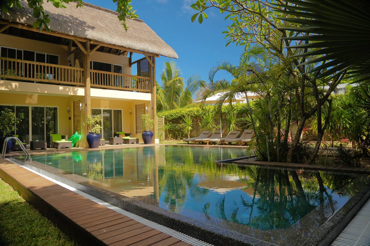 Un espace piscine avec des chaises longues, un bâtiment à toit de chaume et une végétation luxuriante.