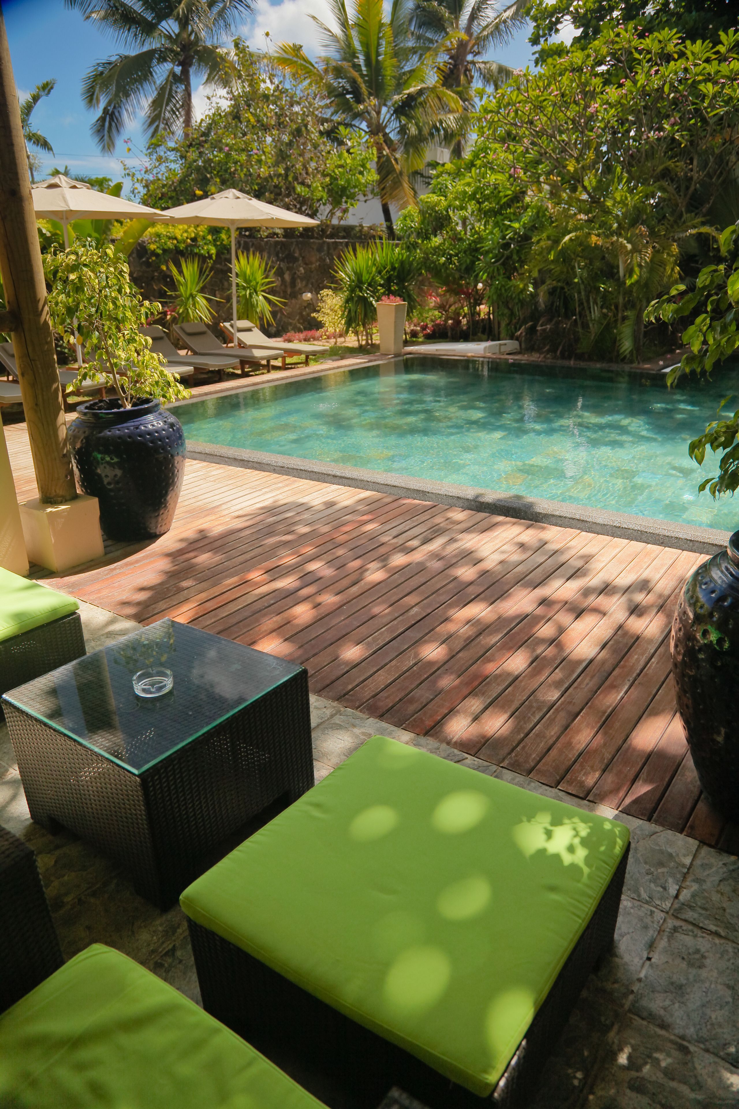Terraza de madera con sillones de descanso y una mesa de vidrio, con vistas a una piscina rodeada de exuberante vegetación.