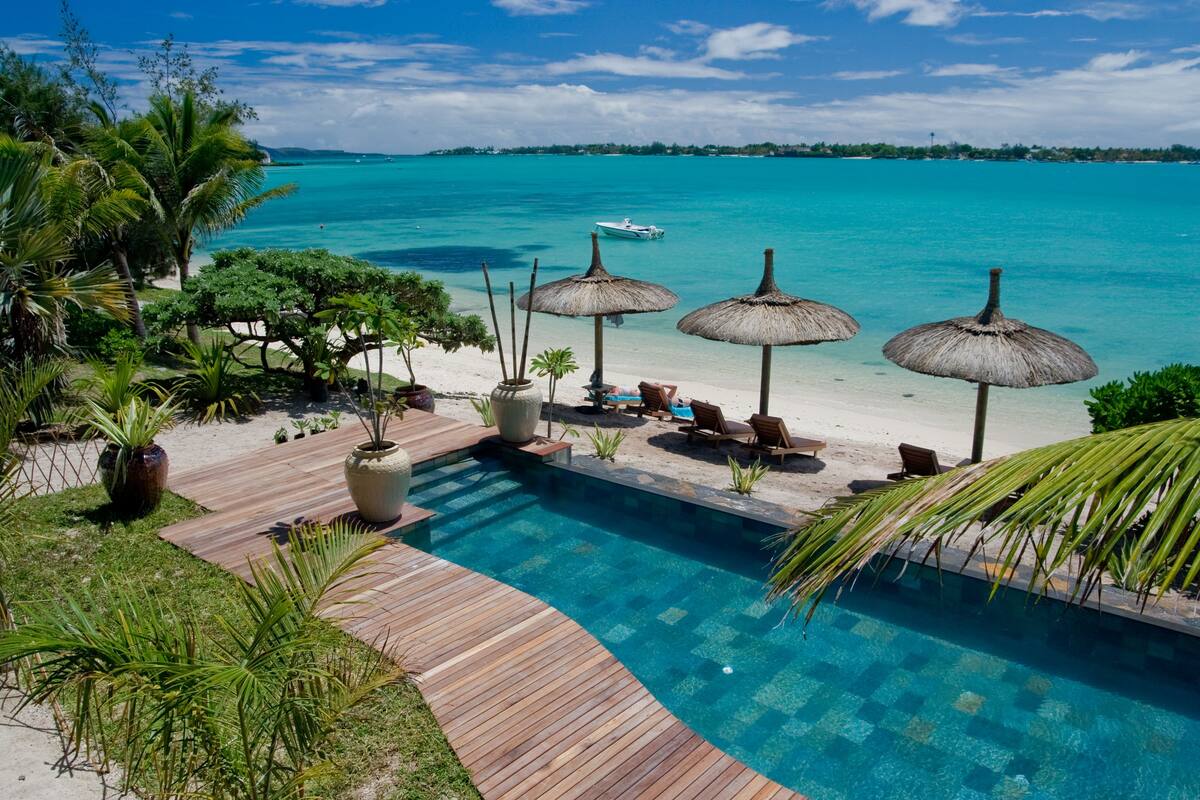 Un espace piscine doté d’une terrasse en bois, de chaises longues et de parasols en chaume.