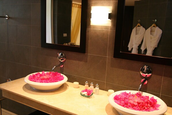 Un baño con dos lavamanos llenos de flores rosas, un espejo y un perchero con batas blancas.