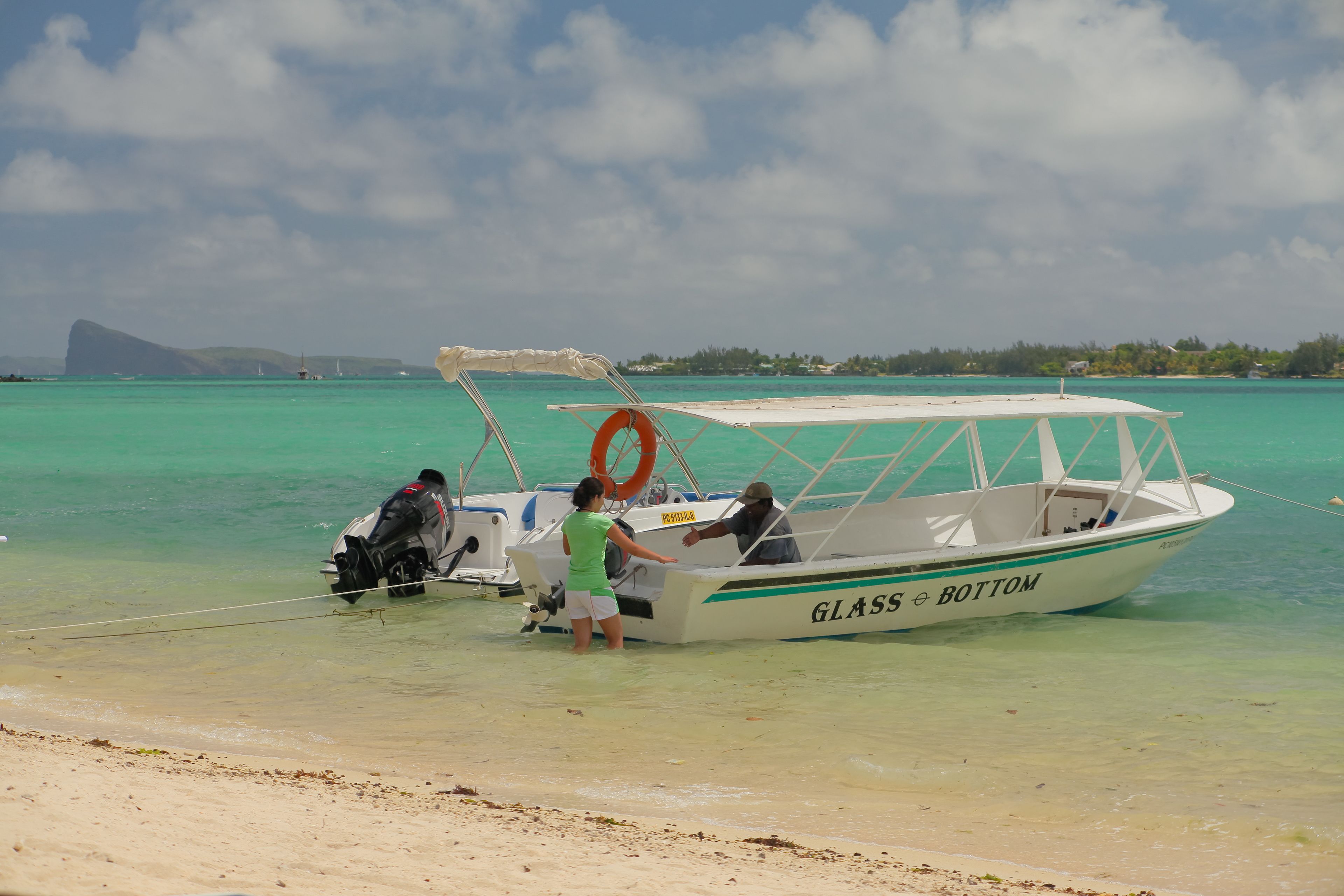 A boat named 'GLASS BOTTOM' on a beach with clear turquoise water and a distant mountain.