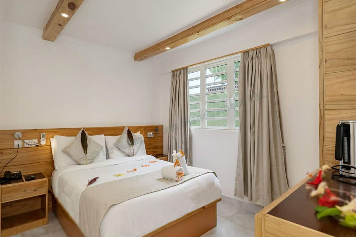 A neatly arranged bedroom with a wooden headboard, a bed with white linens, a bedside table with a remote control, and a window with curtains.