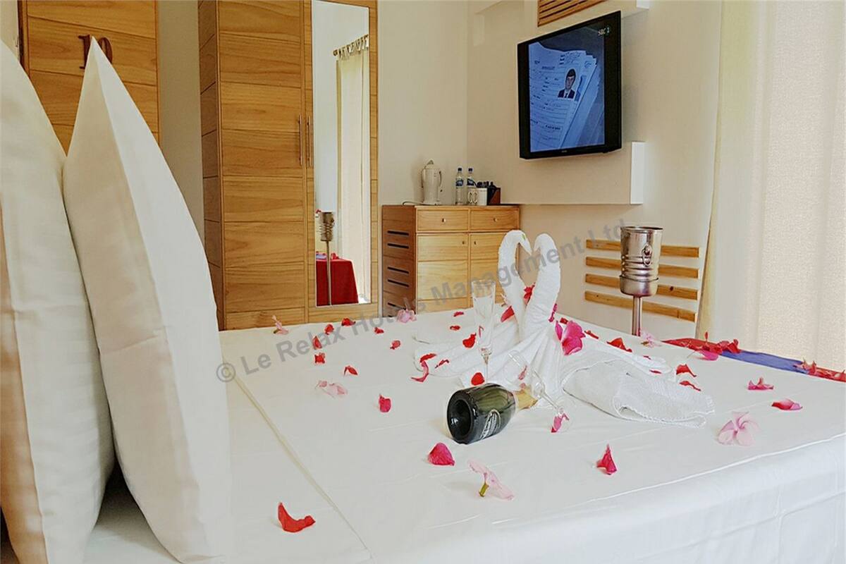 A hotel room with a bed covered in rose petals, a bedside table with a lamp, a television on the wall, and a wooden wardrobe.