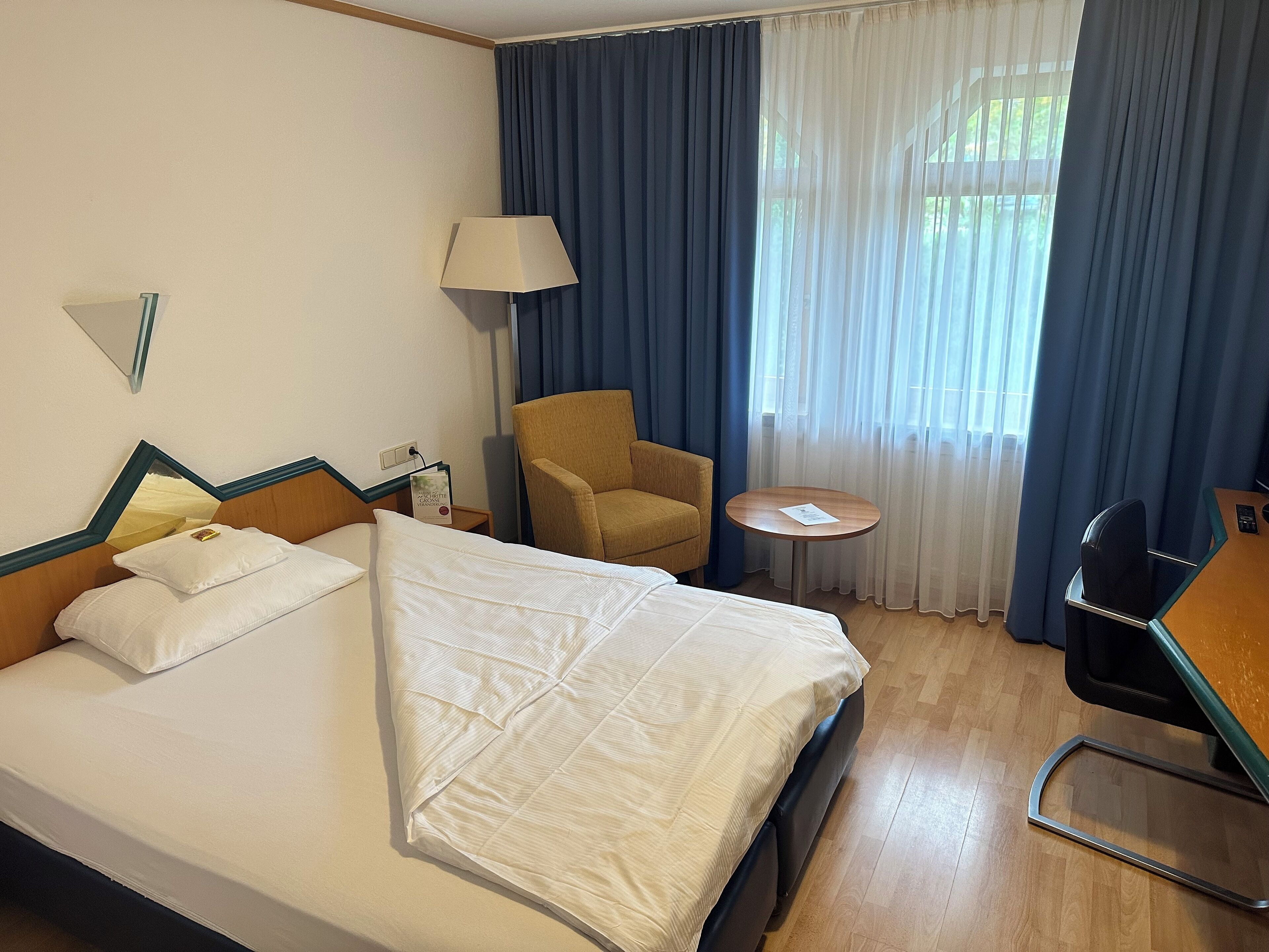 Habitación de hotel con cama, una silla, una mesita, una lámpara y ventana con cortinas.