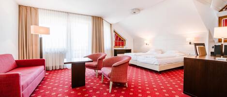 A hotel room with a red carpet, a bed, a desk, a chair, a sofa, and a window with curtains.