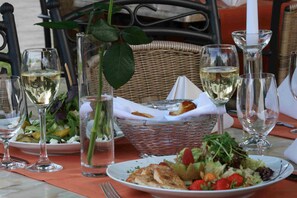 Una mesa de comedor dispuesta con platos de comida, copas de vino blanco y una cesta con pan.