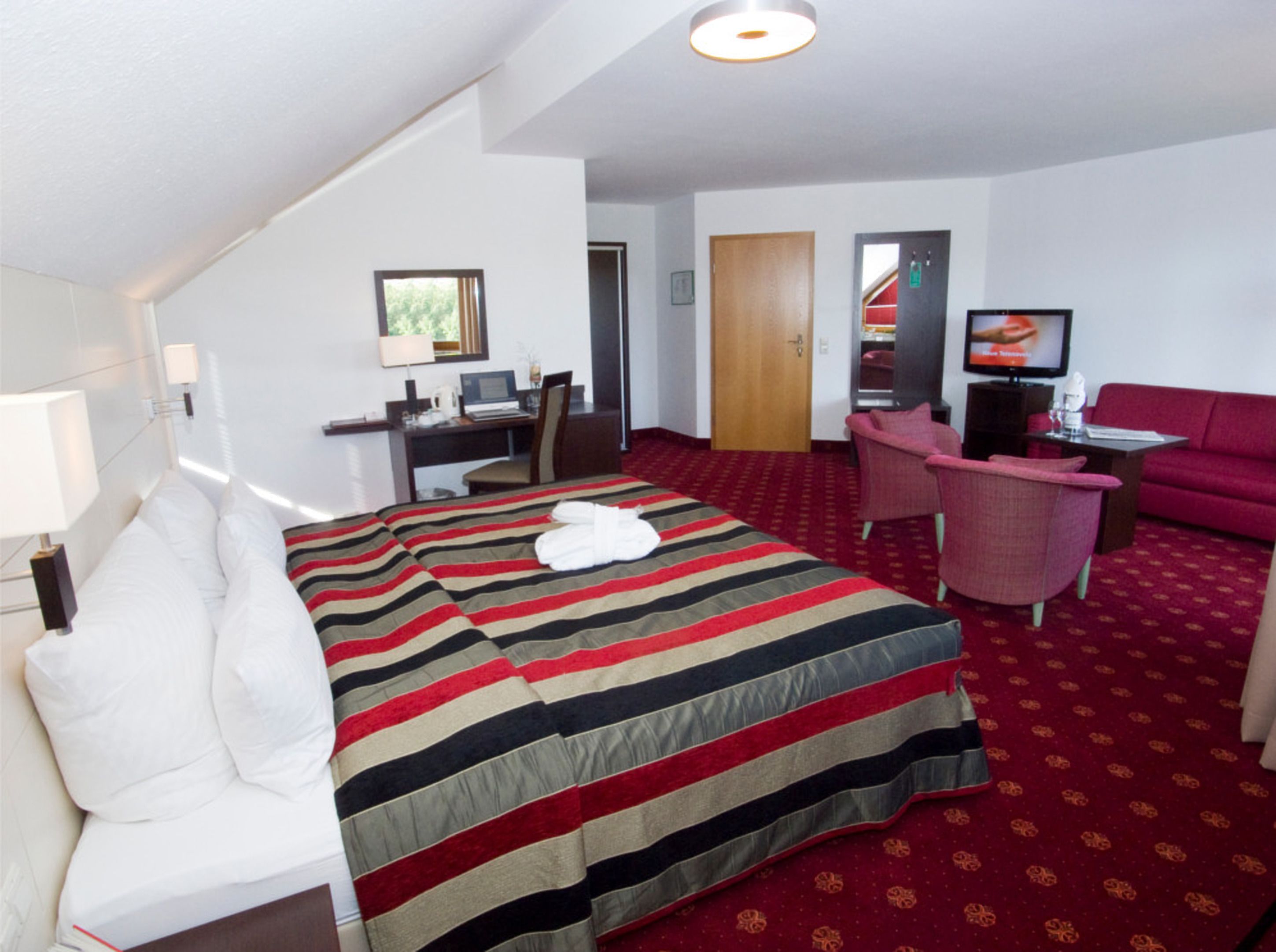 A hotel room with a bed, a desk with a computer, a TV, a red sofa, and a red carpet.