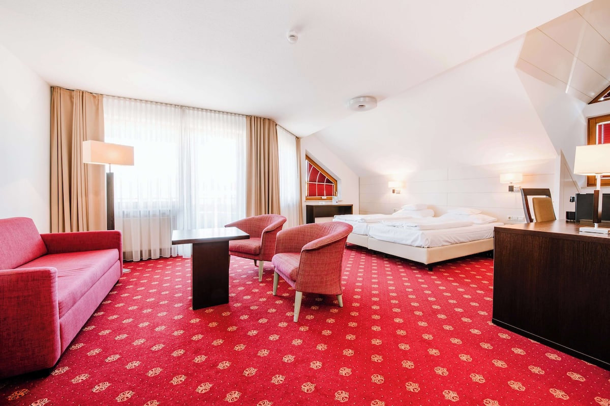 A hotel room with a red carpet, a bed, a desk, a chair, a sofa, and a window with curtains.