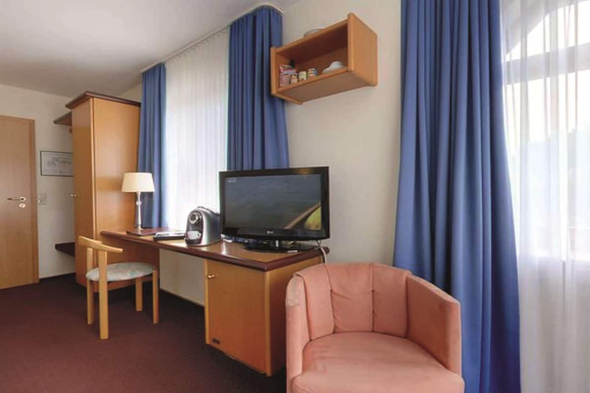 A hotel room with a wooden desk, a television, a chair, a sofa, and a shelf with items.