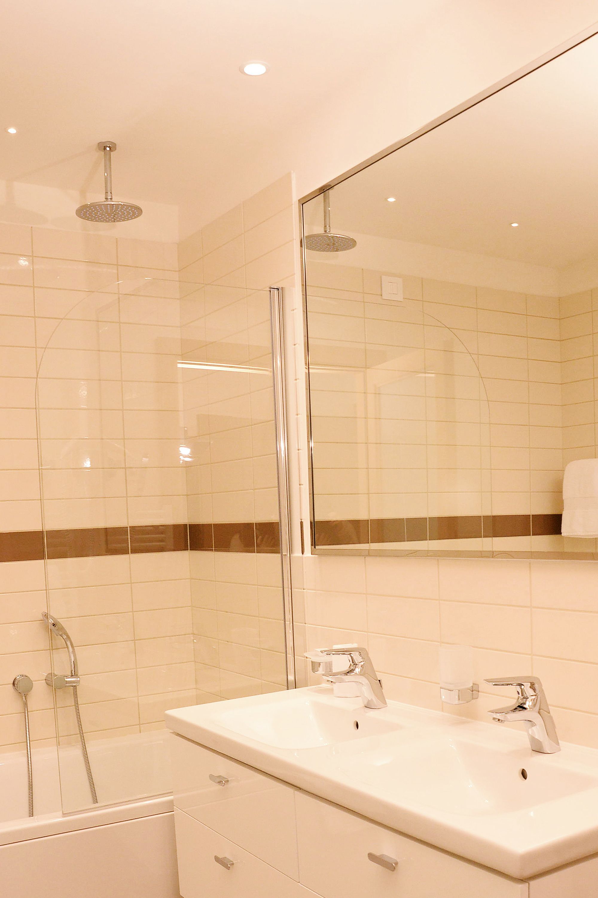 Een moderne badkamer met een douchecabine van glas, een wit wastafelmeubel met twee kranen en een spiegel die de ruimte weerkaatst.