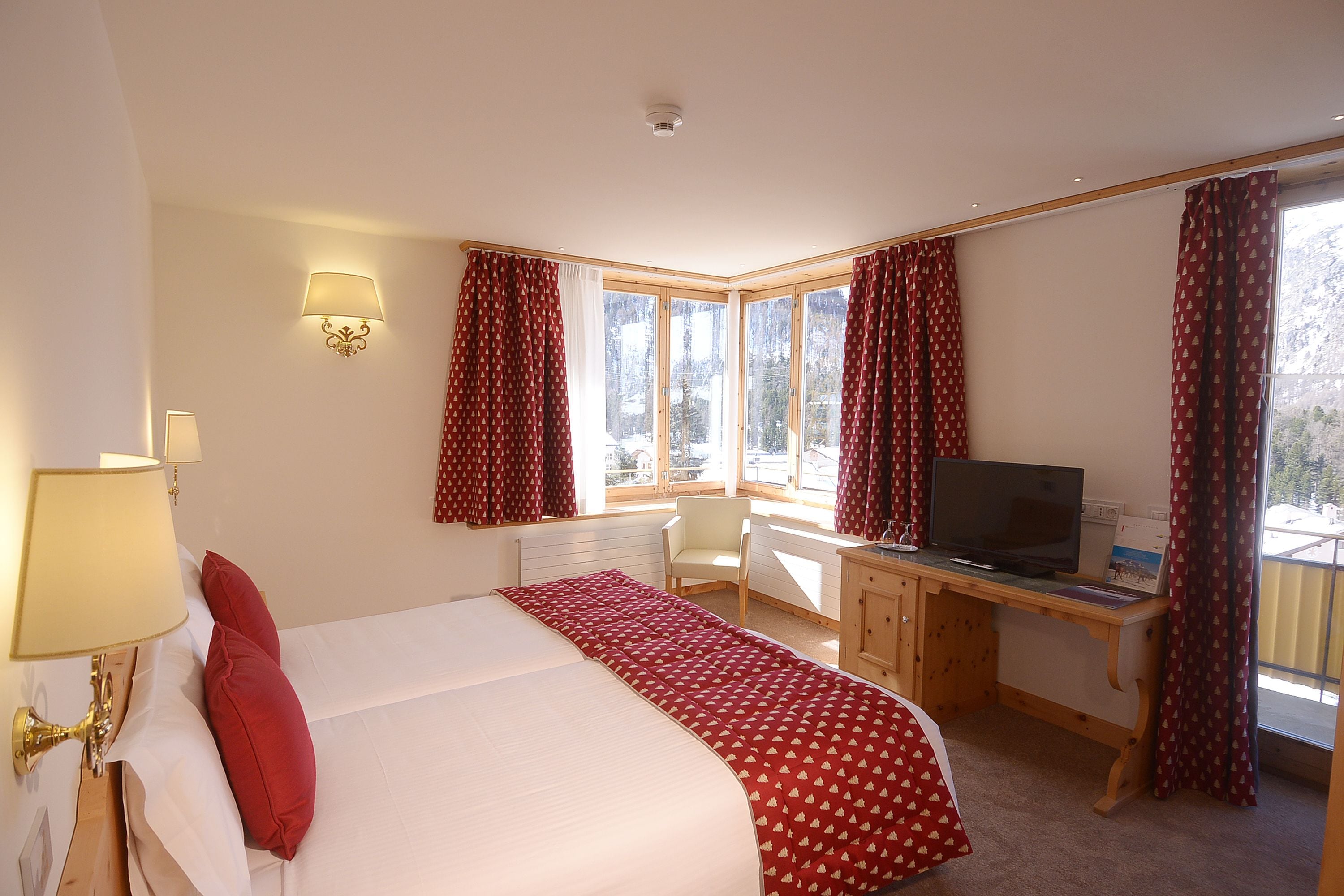 A hotel room with a bed, a desk, a chair, a television, and large windows with curtains.