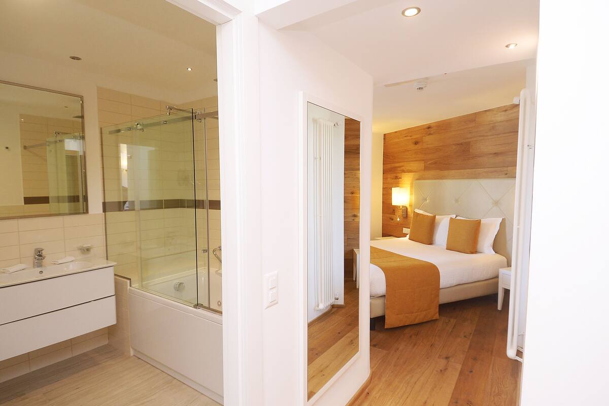 Une chambre d’hôtel moderne dotée d’une douche en verre, d’un grand lit avec une tête de lit en bois et d’une salle de bain séparée avec un meuble-lavabo et un évier.
