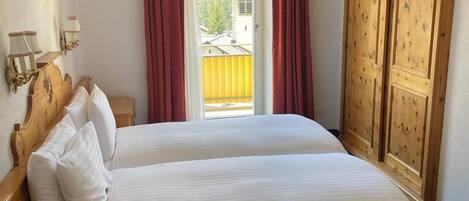 Una cama doble con sábanas blancas, cabecera de madera, mesita de noche con teléfono y ventana con cortinas rojas.