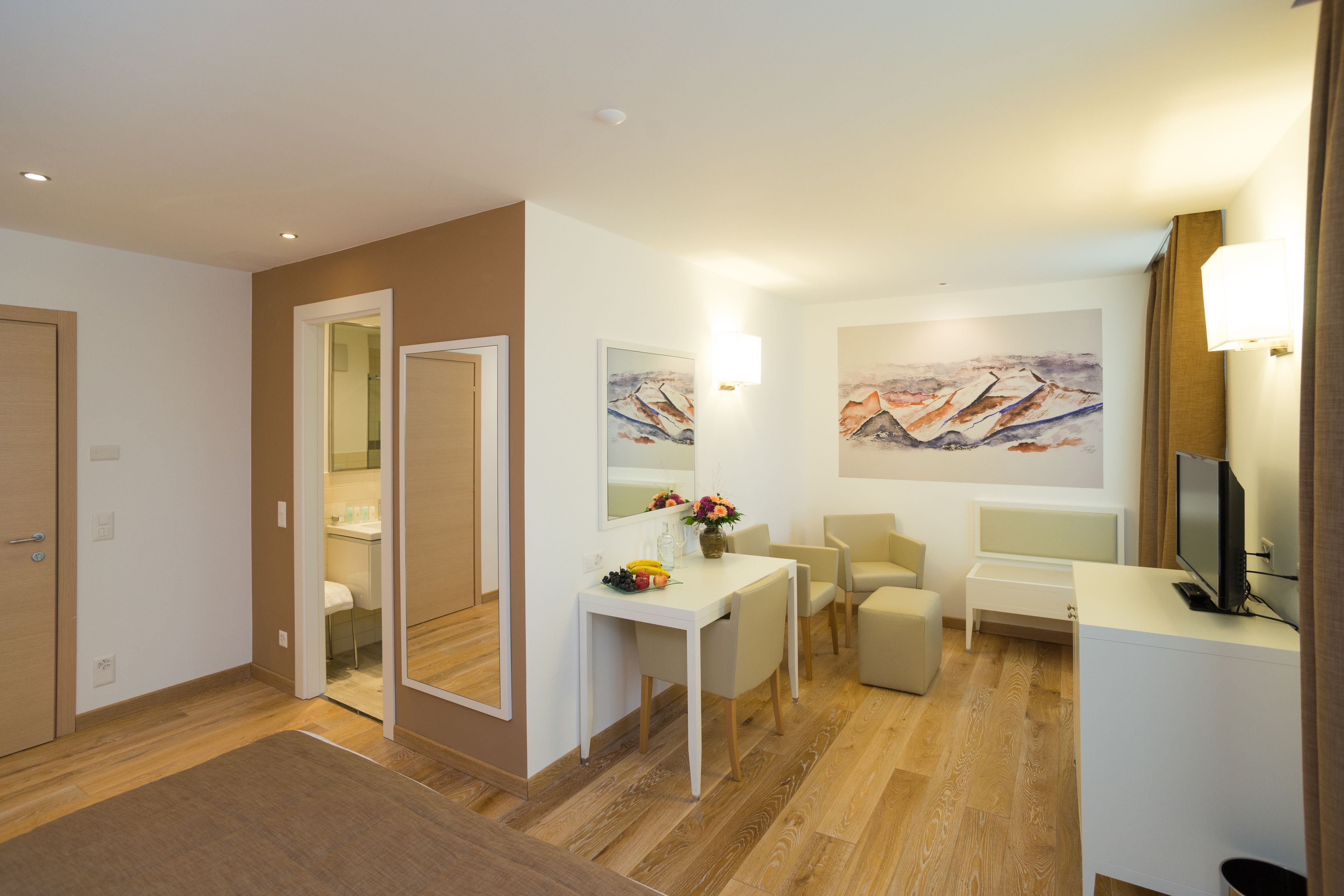 A modern hotel room with a wooden floor, a dining area with a white table and chairs, a TV, and a painting on the wall.