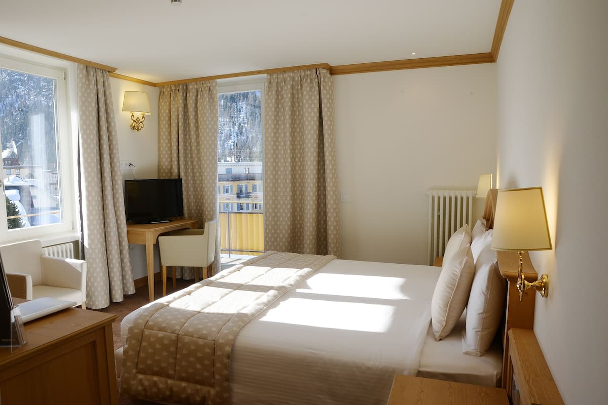 Une chambre d’hôtel avec un lit, un bureau avec un ordinateur, une chaise, une télévision et une fenêtre avec des rideaux.