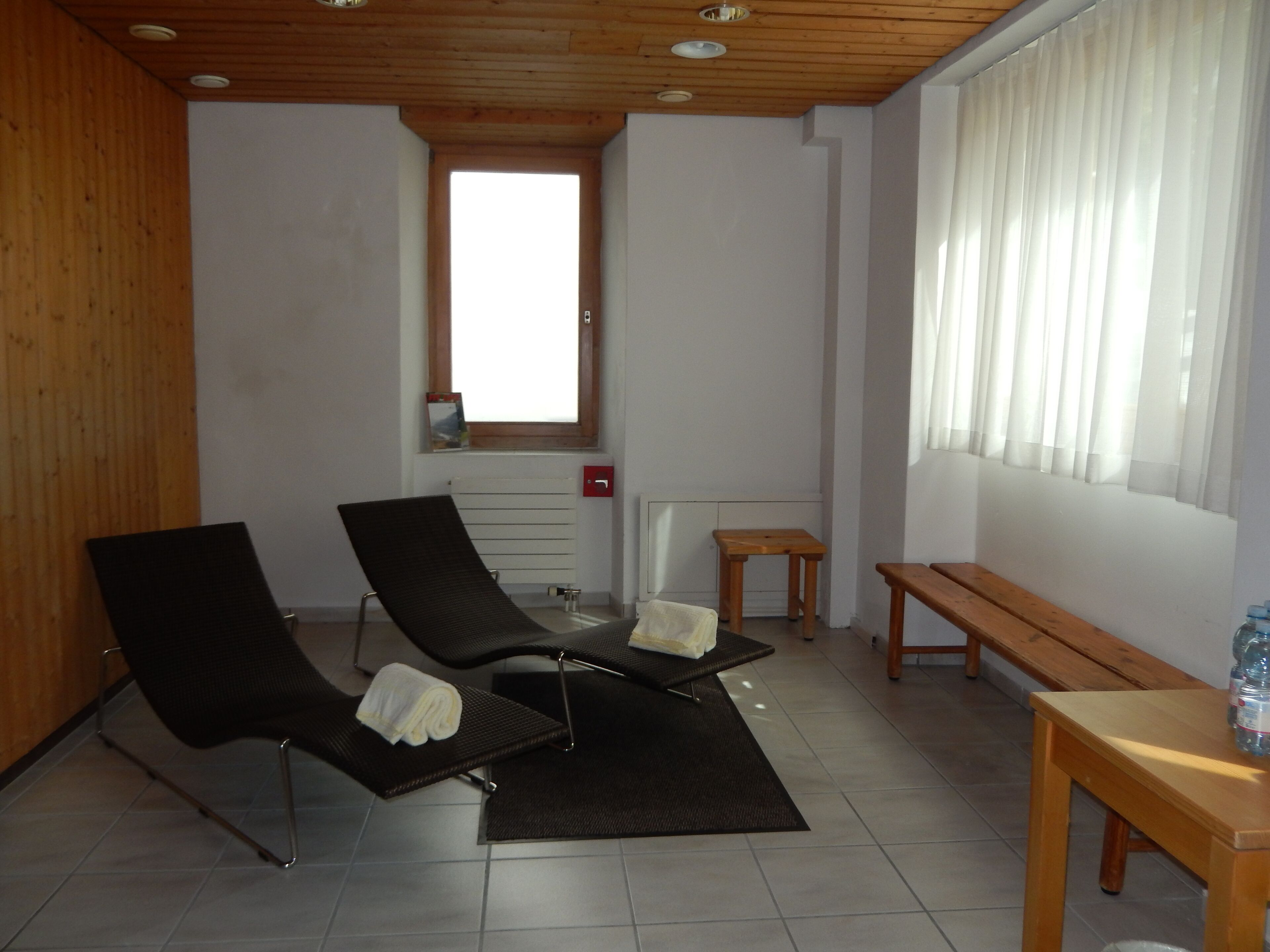Quarto com duas poltronas reclináveis, uma mesa de madeira pequena, um banco e uma janela com cortinas.