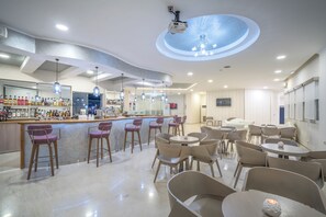 A modern bar area with a counter, bar stools, and a variety of bottles. There are tables and chairs for dining, and a ceiling with a circular light fixture. - Golden Sun (Zakynthos)