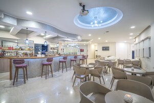 Zona de bar moderna con barra, taburetes y una variedad de botellas. Hay mesas y sillas para comer, y un techo con lámpara circular.