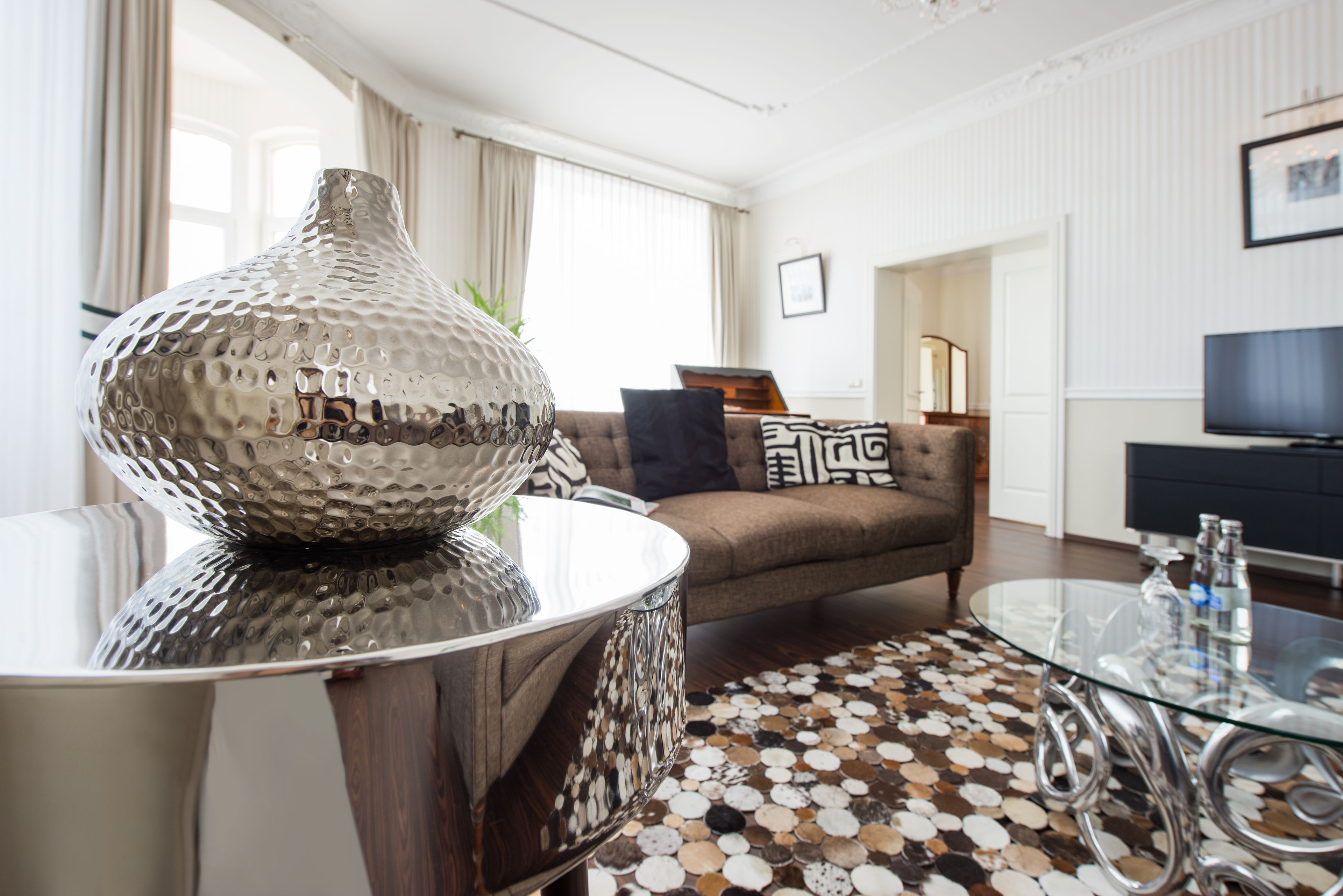 Un salon moderne avec une table basse en verre, un vase décoratif en métal, un tapis à motifs et un canapé marron agrémenté de coussins noirs et blancs.