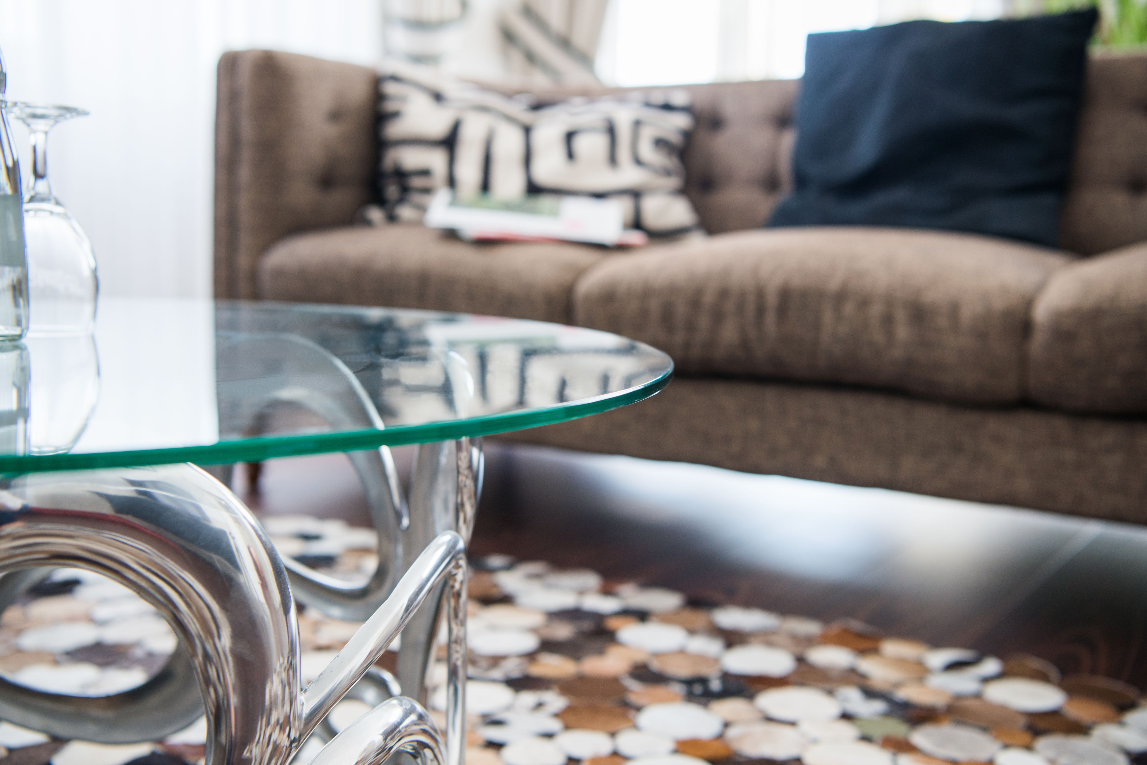 Une table avec un plateau en verre et un pied métallique, un tapis à motifs et un canapé marron agrémenté de coussins, le tout dans un salon.