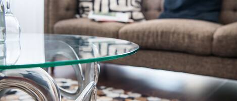 Une table avec un plateau en verre et un pied métallique, un tapis à motifs et un canapé marron agrémenté de coussins, le tout dans un salon.