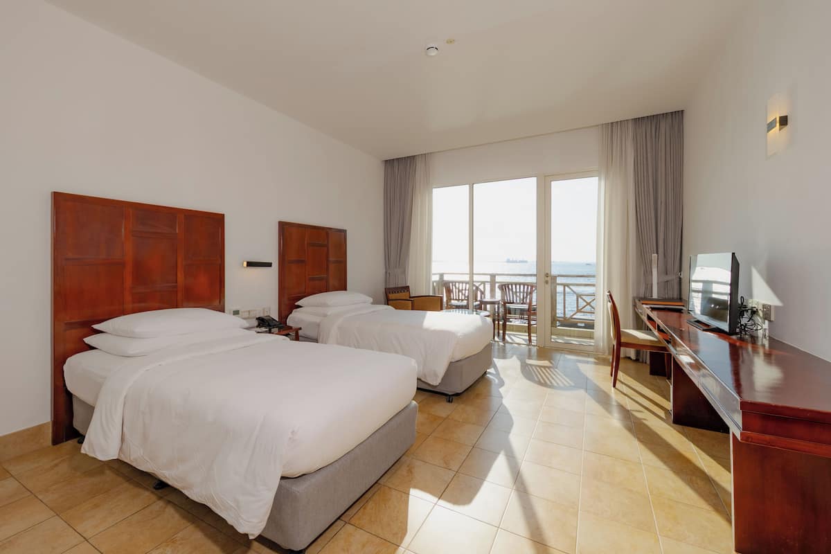 Ein Hotelzimmer mit zwei Betten, einem Schreibtisch, einem Sessel und einem Fernseher. Es gibt einen Balkon mit Meerblick.