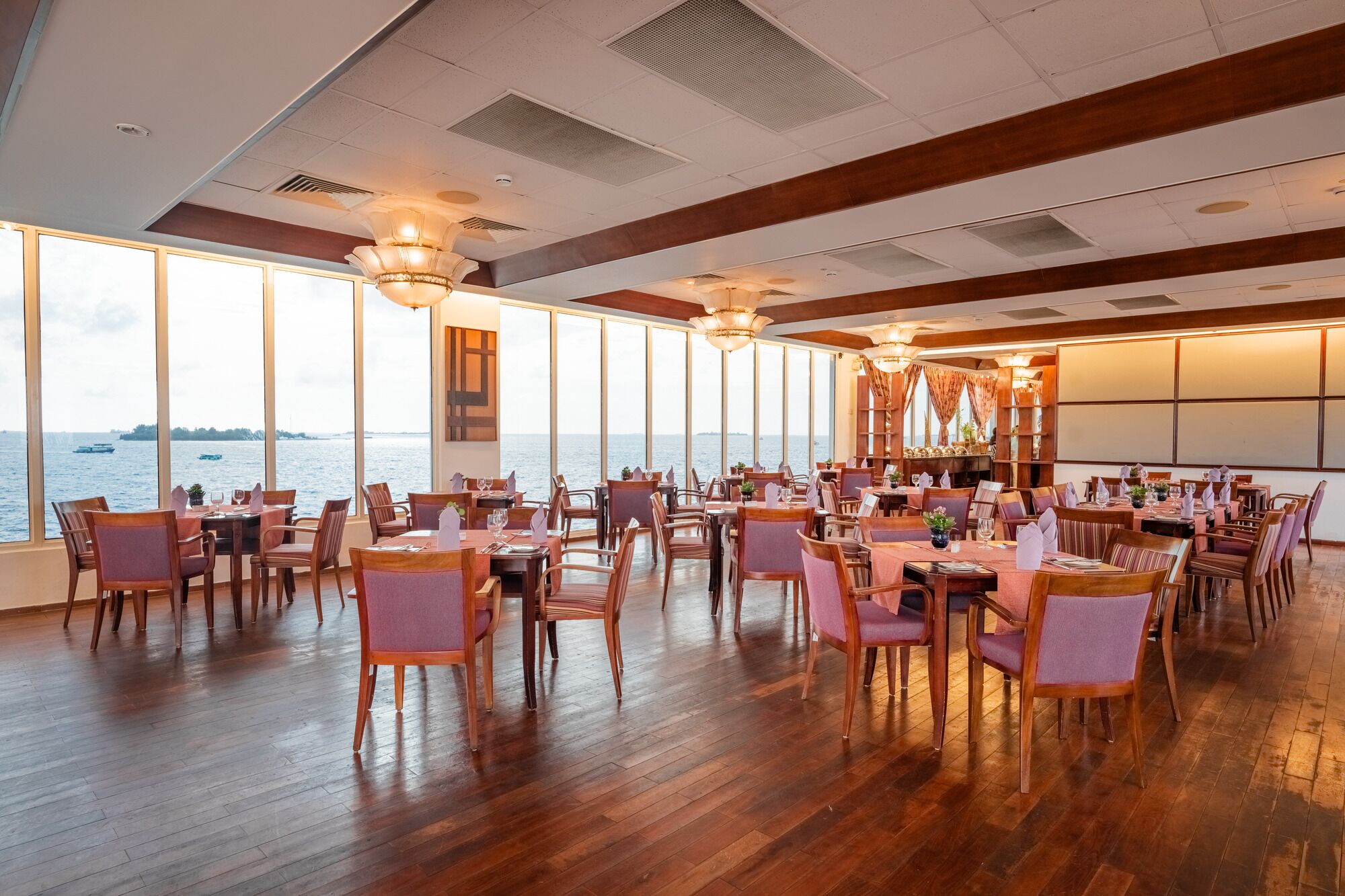 Uma sala de jantar ampla com piso de madeira e janelas grandes com vista para o mar.