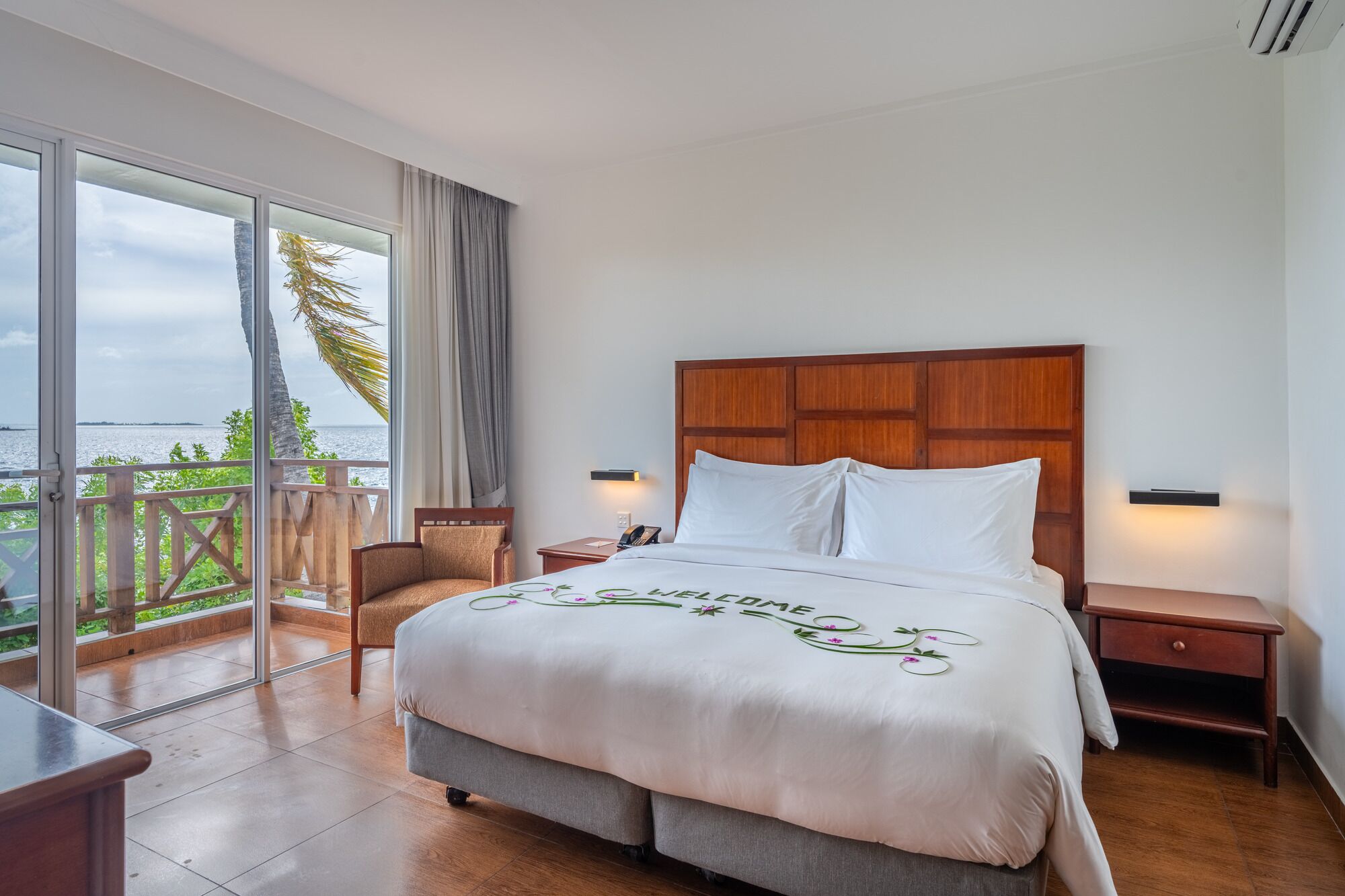Ein Hotelzimmer mit einem großen Bett, einem Holz-Kopfteil, einem Nachttisch, einem Sessel und einem Balkon mit Meerblick.