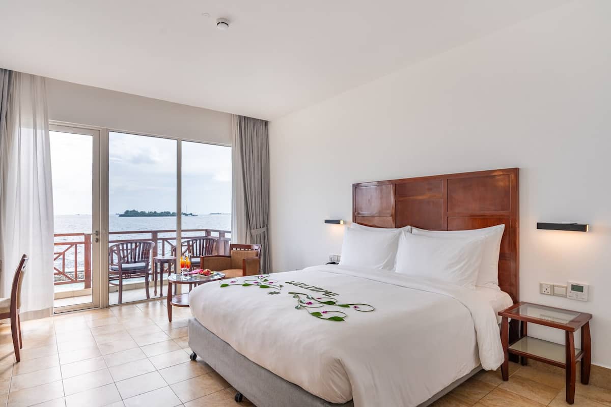 Ein Hotelzimmer mit einem großen Bett, Meerblick, einem Balkon mit Stühlen und einem kleinen Tisch mit Getränken.