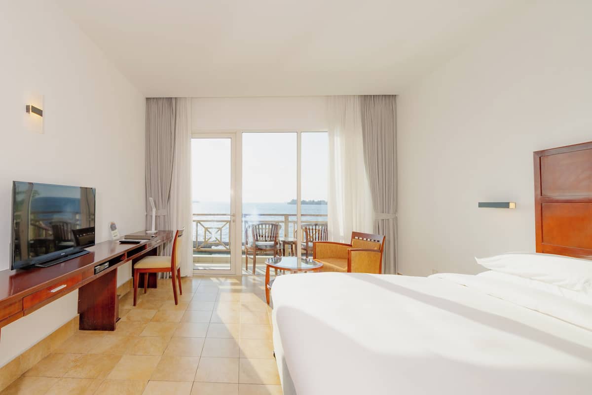 Ein Hotelzimmer mit Bett, Schreibtisch mit Fernseher, Sessel, kleinem Tisch und einem Balkon mit Meerblick.