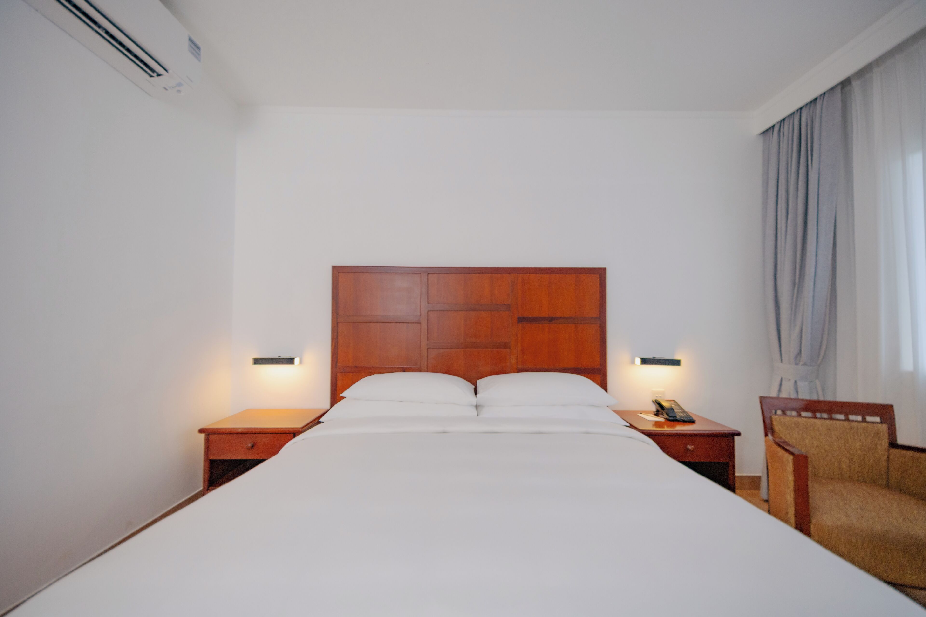 A hotel room with a wooden headboard, a bed with white linens, two bedside tables with lamps, and a chair.