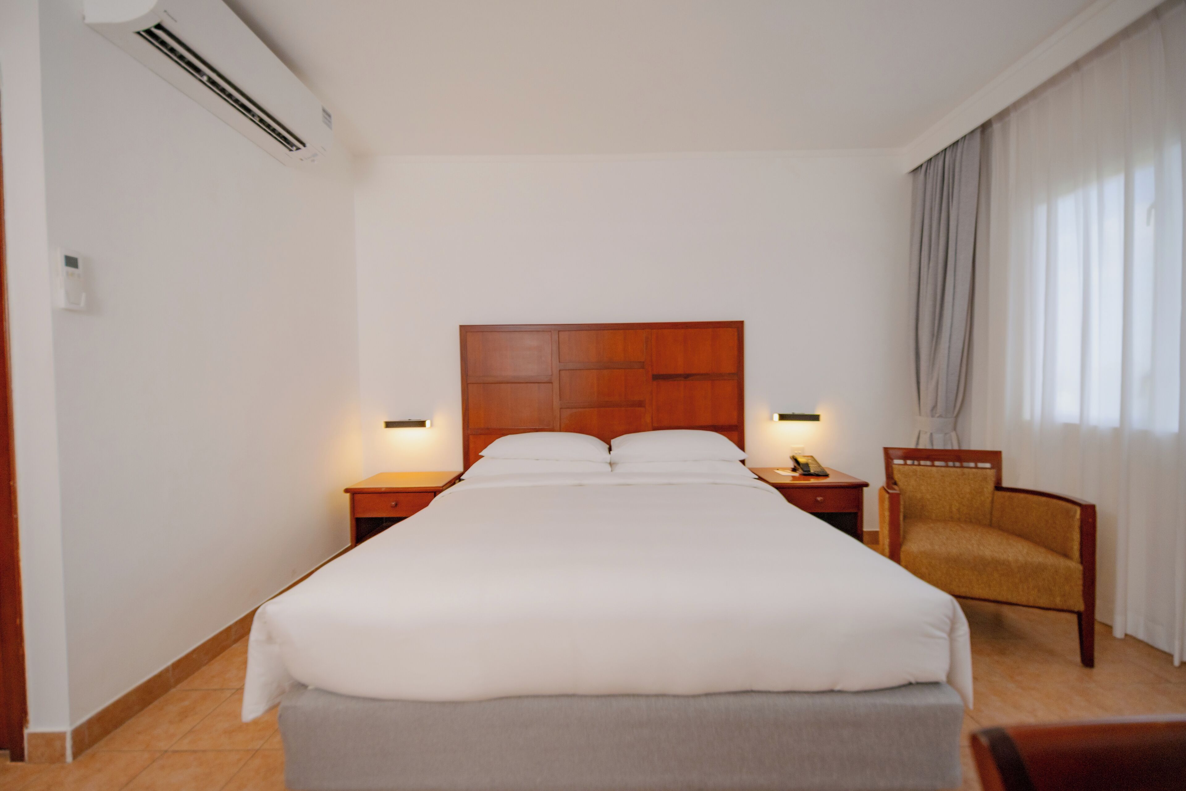 Een hotelkamer met een groot bed, een houten hoofdbord, bedlampjes, een stoel en een raam met gordijnen.