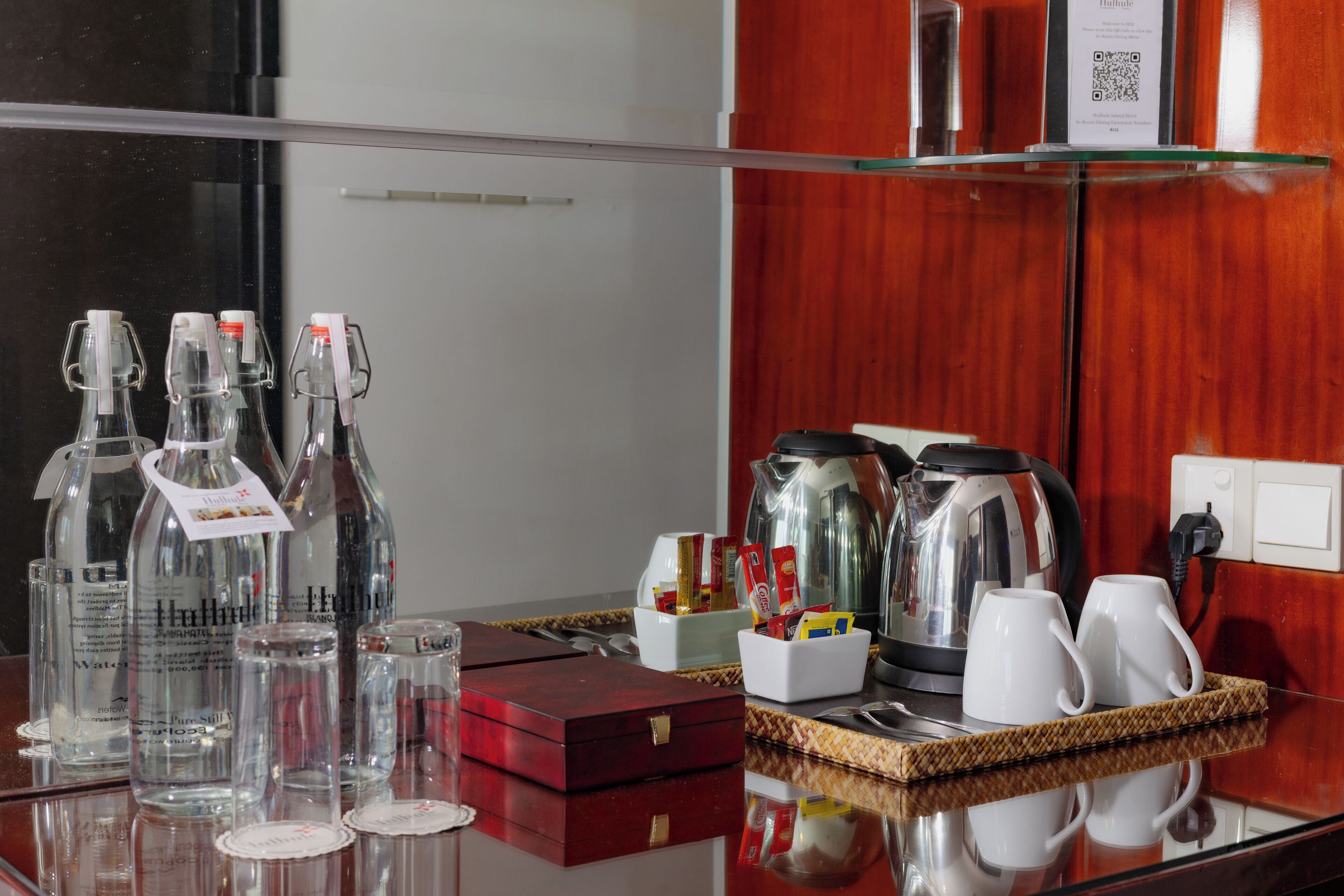 A hotel room's breakfast area with a glass-top table, clear bottles, glasses, a coffee maker, and a tray with cups and condiments.