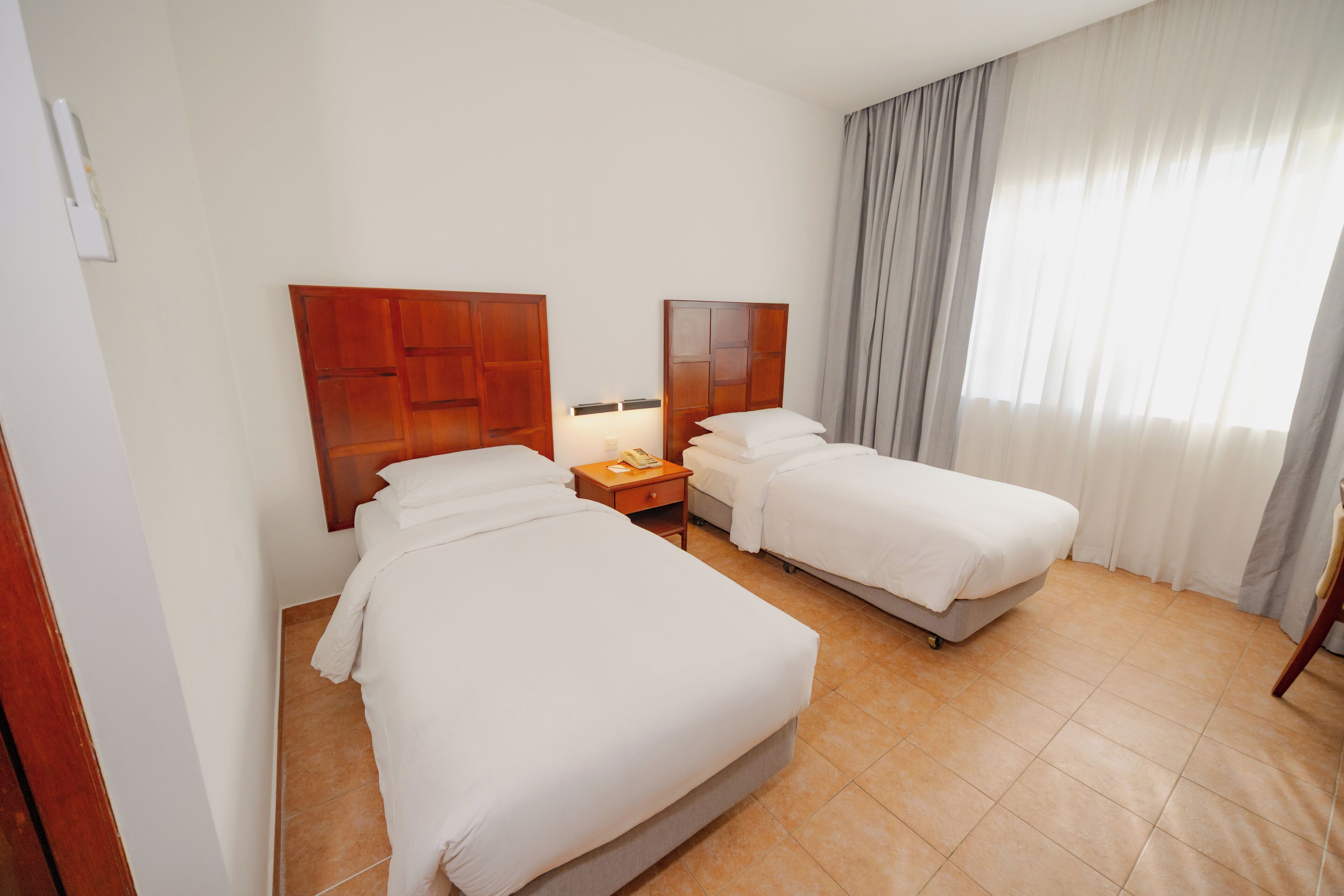 Habitación de hotel con dos camas, una mesita de noche, una lámpara y un ventanal con cortinas.