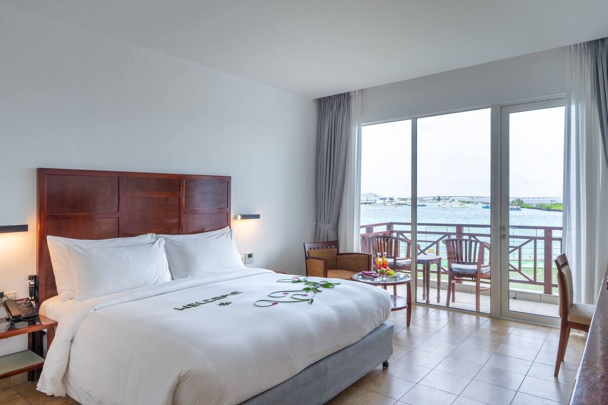 Ein Hotelzimmer mit einem großen Bett, einem kleinen Tisch mit Getränken, einem Sessel und einem Balkon mit Blick auf das Wasser.
