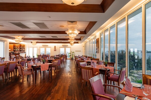 A spacious dining area with wooden flooring and large windows offering a city view. Tables are set with chairs and centerpieces. - Hulhule Island Hotel- Private Beach Access & Airport Pick-up and Drop (Hulhulé)