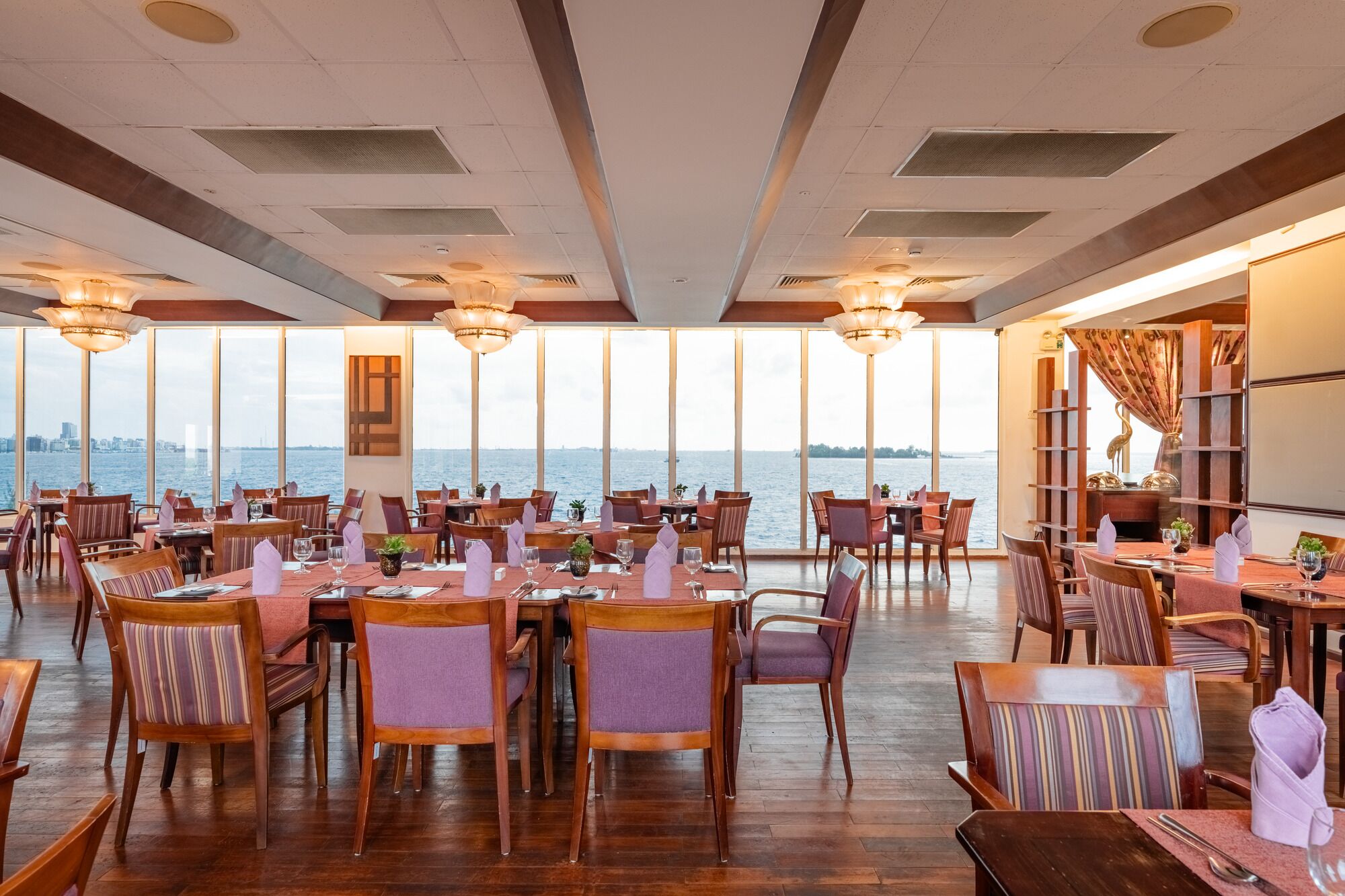 A restaurant with multiple tables and chairs, large windows with a view of the water, and a modern ceiling design.