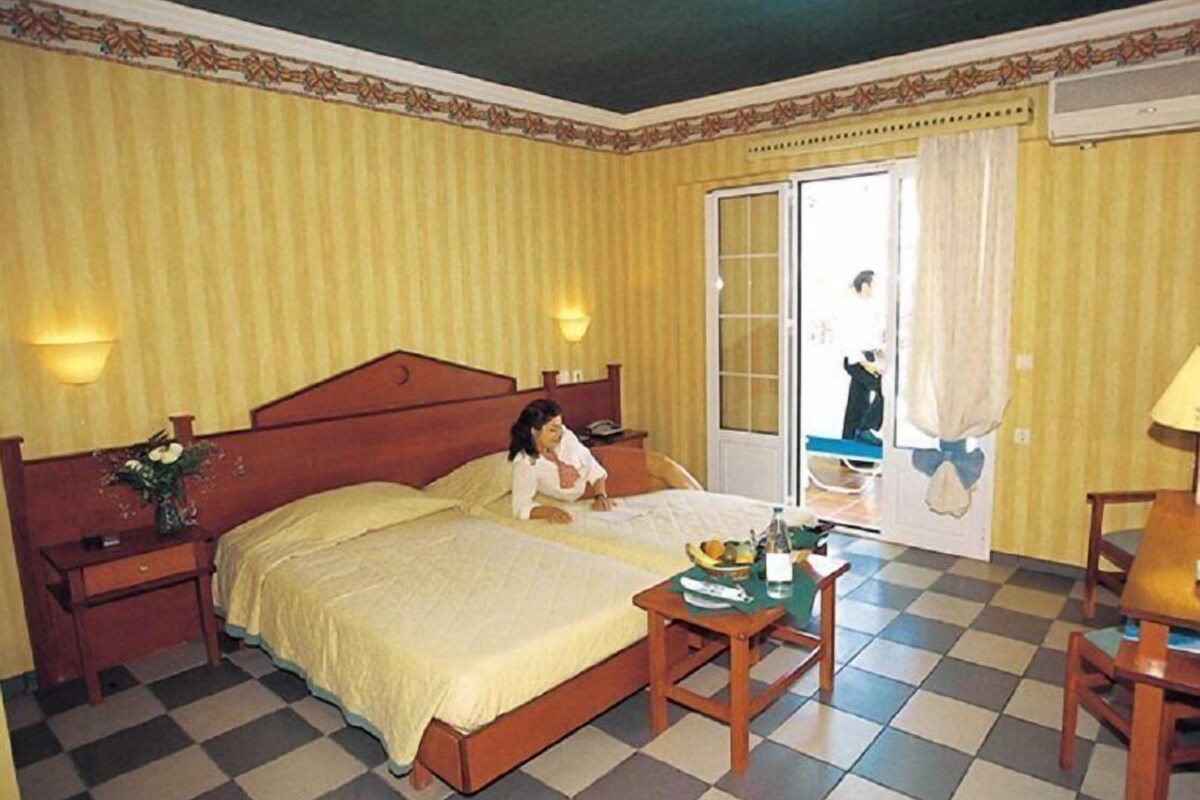 A hotel room with a bed, a small table with a bottle and fruit, a chair, and a view of the outside.