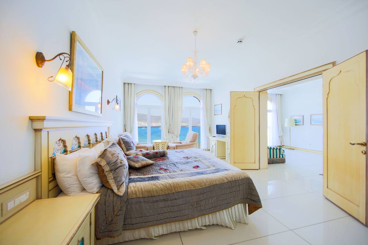 A spacious bedroom with a large bed, a sitting area with a view, and a door leading to another room.