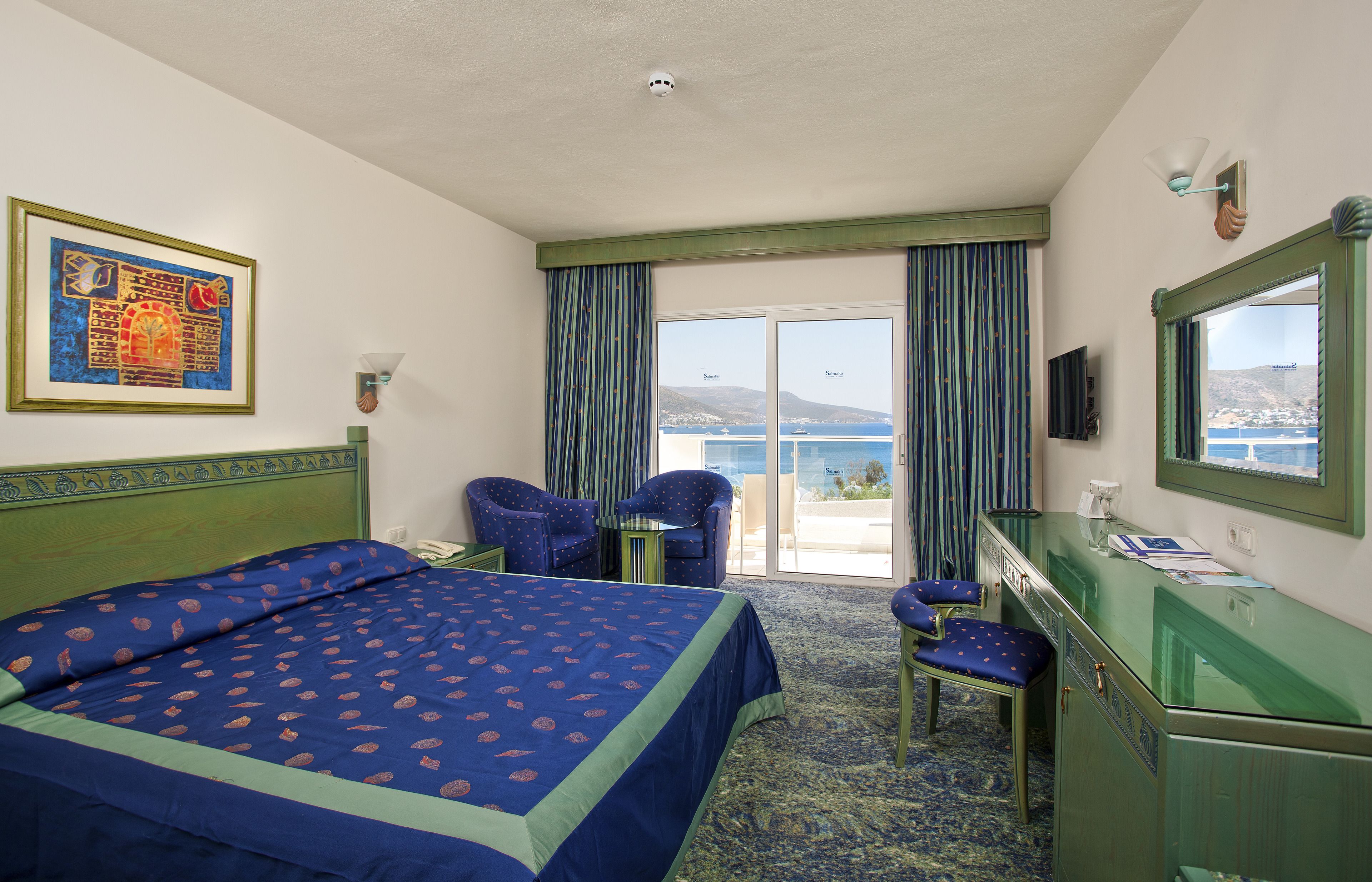 Habitación de hotel con cama, dos sillas, escritorio, espejo y vista al mar.