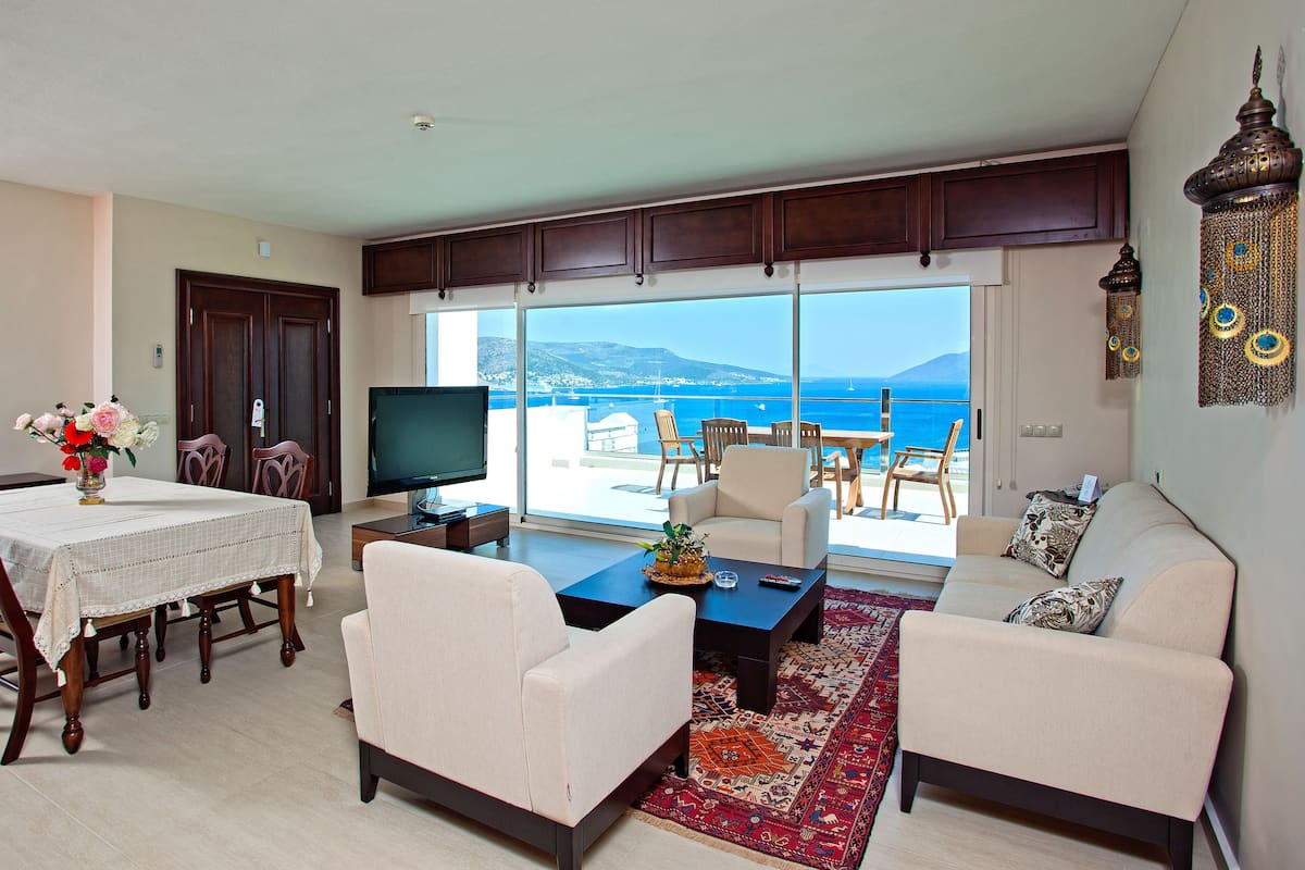 A living room with a TV, dining table, and chairs, a sofa, and a view of the sea.