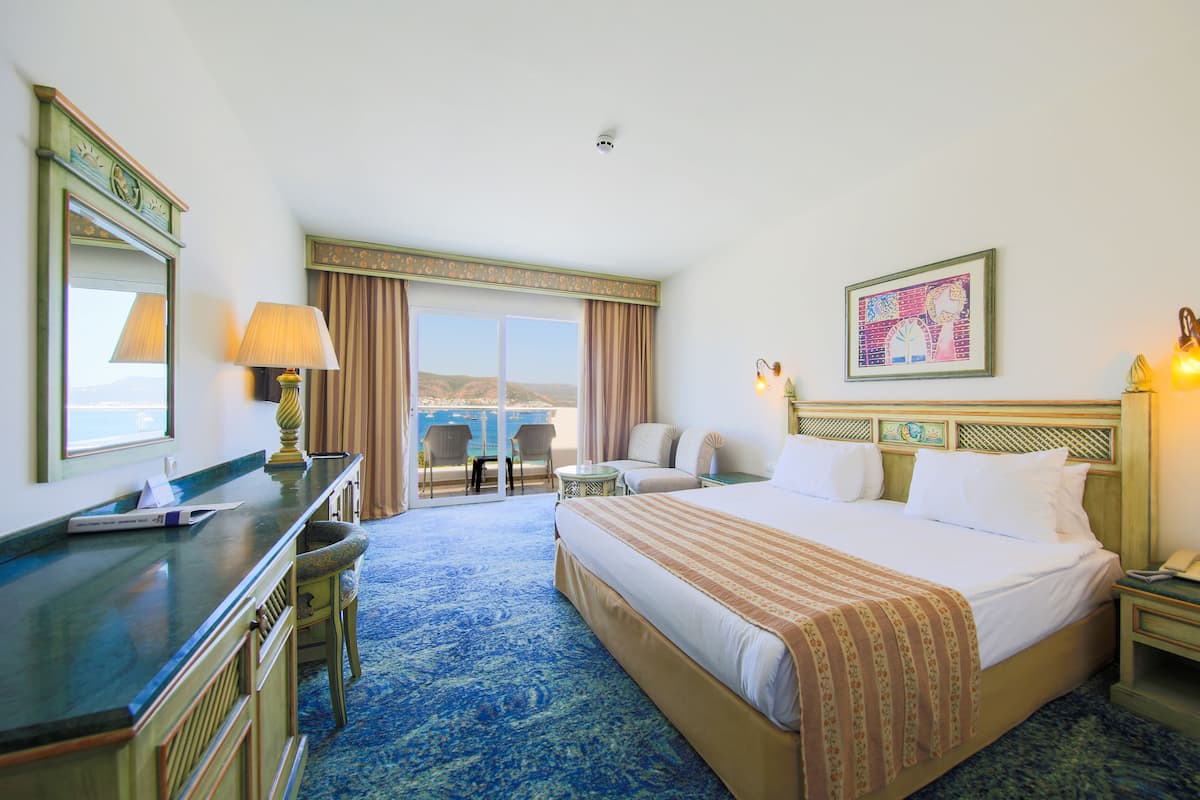 A hotel room with a large bed, a desk with a lamp, a chair, a small table, and a view of the outdoors.