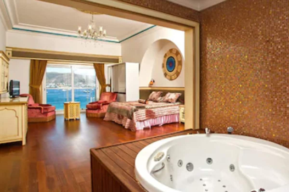 A spacious bedroom with a large bed, a sitting area with a chair, a large bathtub, and a view of the sea through a large window.