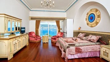 Un dormitorio espacioso con una cama grande, un escritorio con televisor, dos sillones y vistas al mar a través de un ventanal amplio.