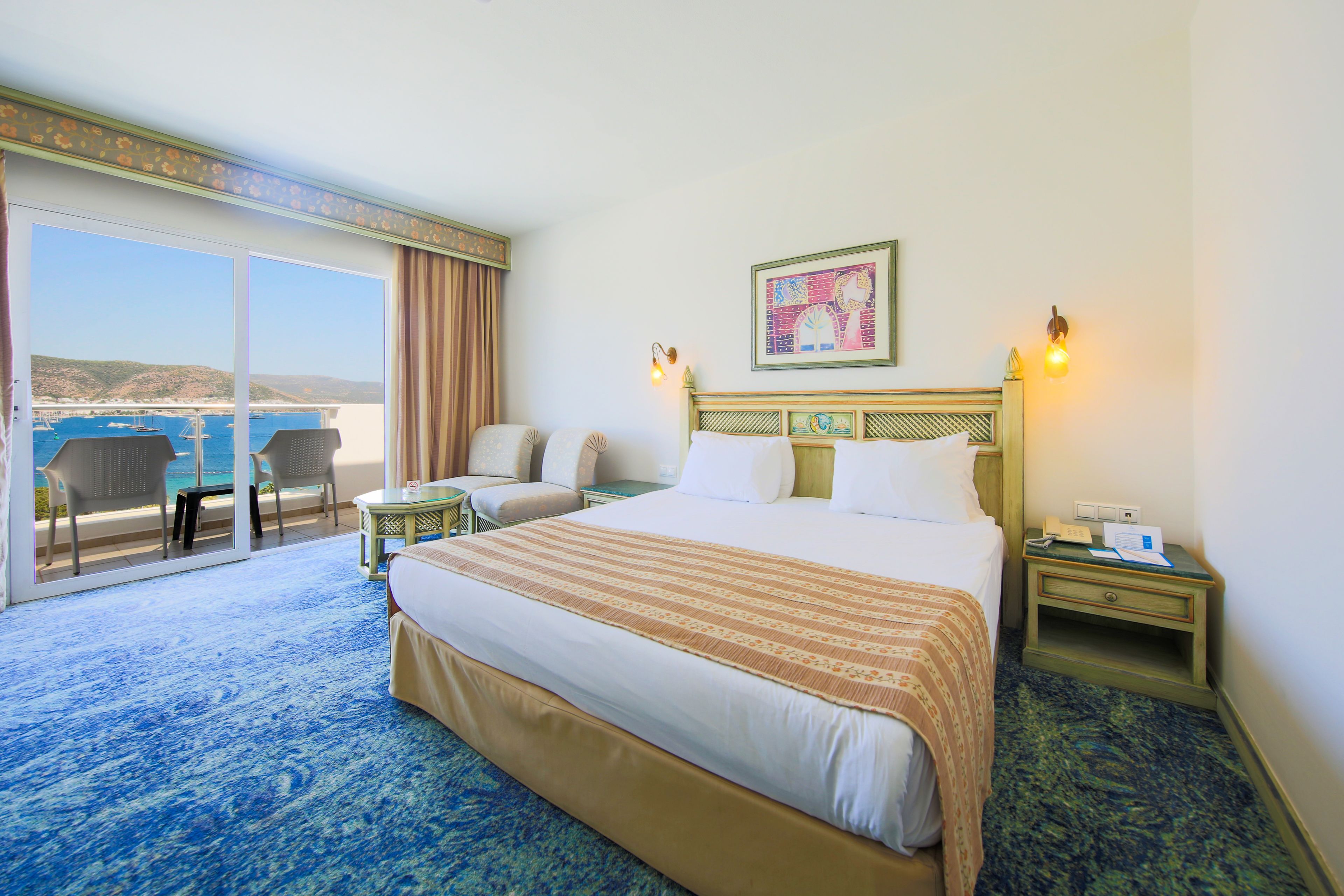 A hotel room with a large bed, a balcony with chairs, a small table, and a nightstand with a lamp.