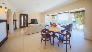 A living room with a TV, sofa, dining table, and chairs. There is a balcony with a view of the sea.