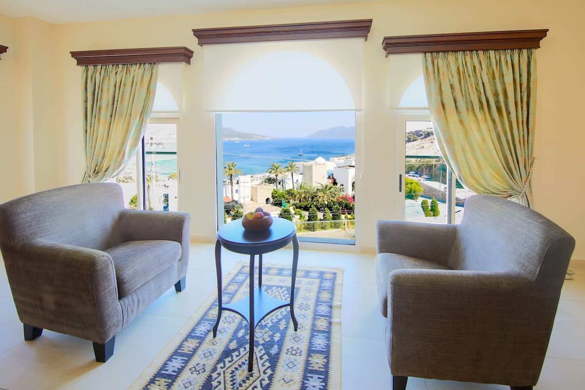 A room with two armchairs, a small table, and a view of the sea and buildings.