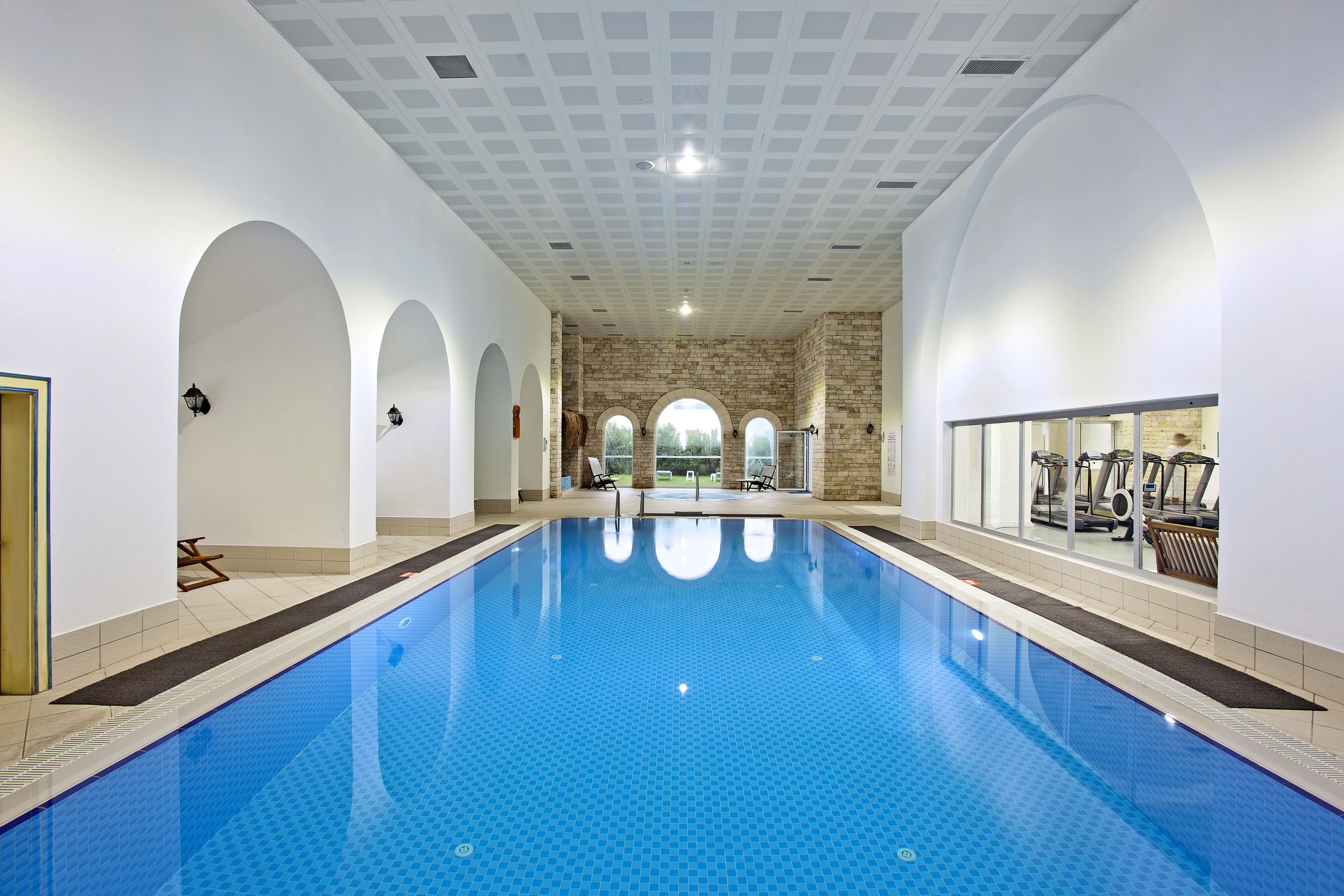 An indoor swimming pool with clear blue water, surrounded by white walls and arches, and a brick wall in the background.